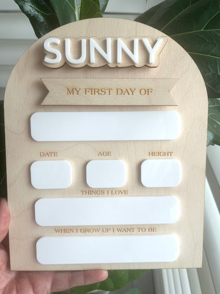 A wooden sign with the words 'SUNNY' at the top and spaces for personalization such as date, age, and a message. It is designed for writing with whiteboard markers for tracking a child's first day of school.