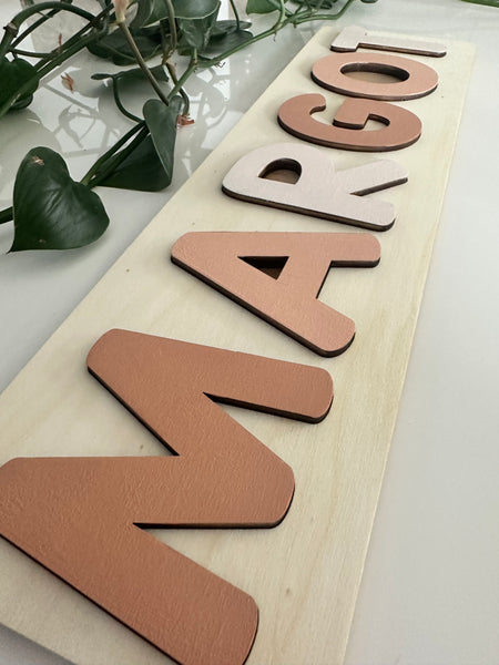 A personalized wooden name sign spelling 'MARGOT' in raised letters mounted on a light wood panel, styled with green leaves on a white surface.