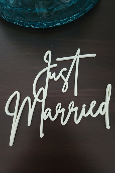 White script cake topper reading 'Just Married' placed on a light wood surface with a teal glass plate partially visible at the top.
