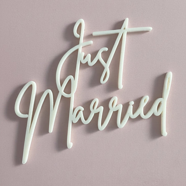 White script acrylic cake topper reading 'Just Married' displayed on a dusty pink rectangular board.