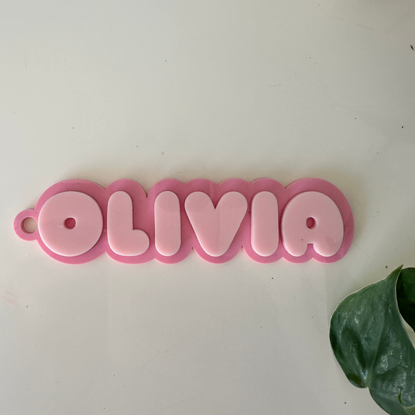 A pink personalized name sign spelling 'OLIVIA' in raised bubble letters, displayed on a light off-white surface with a green leaf in the corner.