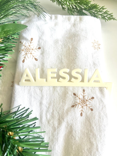 White fabric with gold snowflake patterns and 'ALESSIA' name, surrounded by greenery