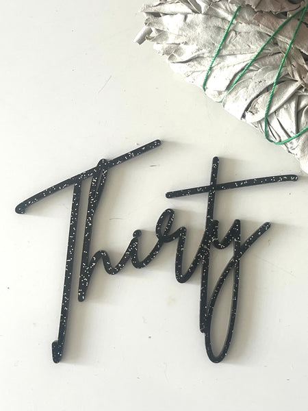 Black glittery 'Thirty' cake topper on a white background with silver leaves.