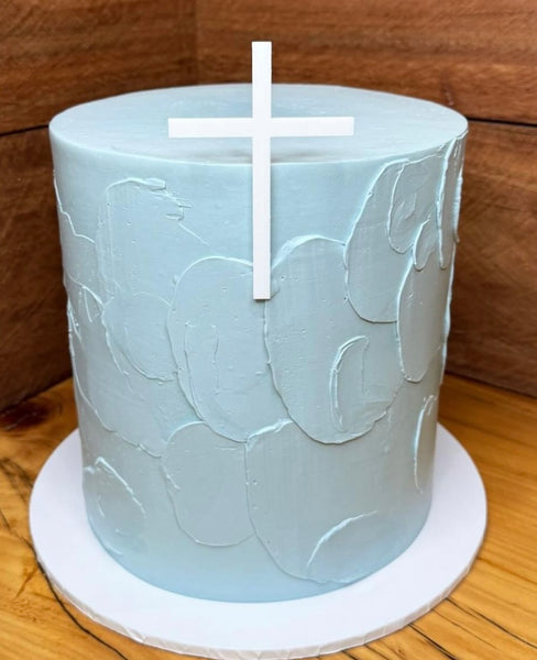 A cake with a gold acrylic cross charm and a personalized name charm in black lettering on top of a white frosting covered cake with gold accents.