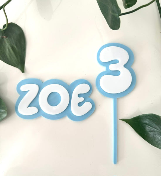 Cake topper with 'ZOE' and number '3' on a white background with green leaves.