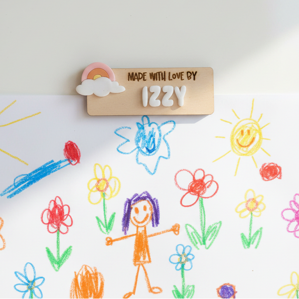 A white square art print featuring a colorful children's crayon drawing of flowers, suns, and a stick figure, hung on a light wall with a wooden sign that reads 'MADE WITH LOVE BY IZZY'.